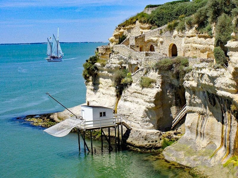 Cabane de pêcheur sur pilotis avec filet suspendu au pied d'une falaise calcaire face à un voilier en mer.