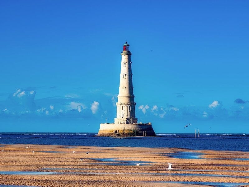 Phare blanc sur une base rocheuse entourée d'eau avec un ciel bleu et quelques mouettes sur le sable.