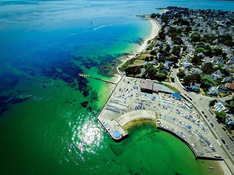 Vue aérienne d'une plage avec un port, des bâtiments et une mer turquoise sous un ciel clair.