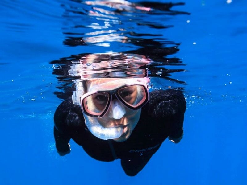 Plongeur avec masque et tuba regardant vers la caméra sous l'eau, reflet à la surface calme.