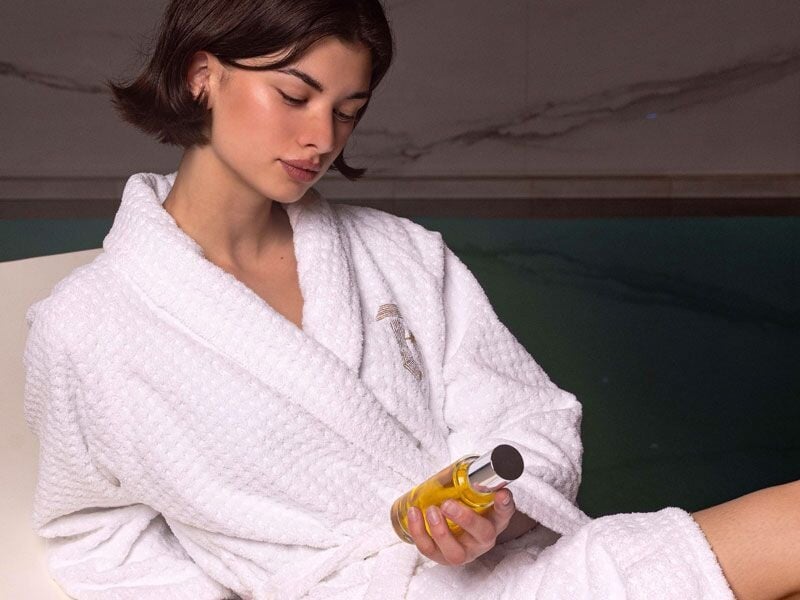 Jeune femme en peignoir blanc tenant une bouteille d'huile de soin près d'une piscine.