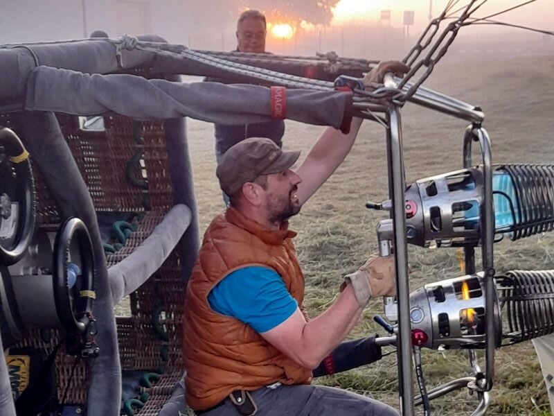 Un homme ajuste le brûleur d'une montgolfière au lever du jour dans un champ brumeux, prêt pour le décollage.