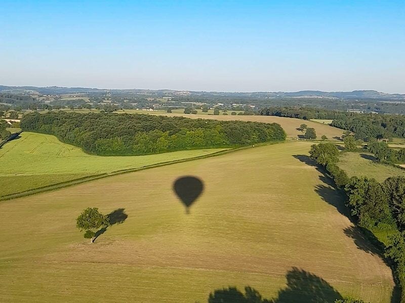 Ombre d'une montgolfière projetée sur un champ vert entouré d'arbres sous un ciel bleu clair.