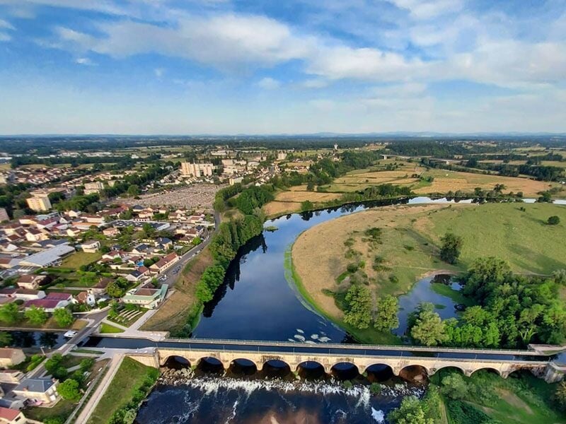 Photo prise en hauteur montrant un pont ancien sur une rivière, avec une ville et des arbres autour.