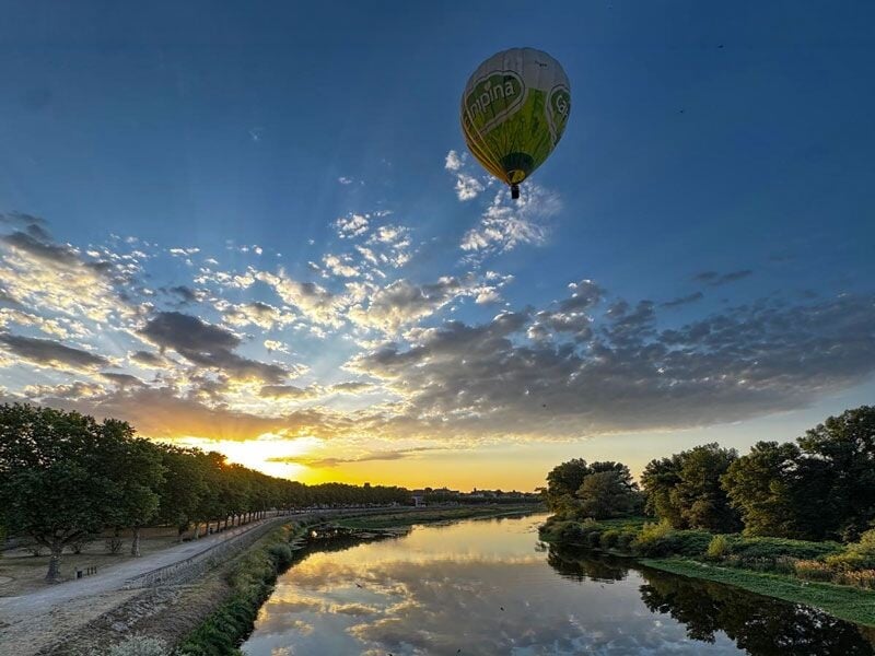 Montgolfière flottant au-dessus d'une rivière au coucher du soleil, avec un ciel nuageux et des arbres en bordure.