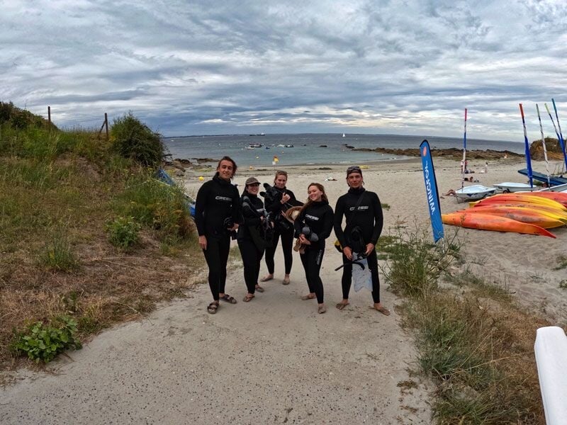 Cinq plongeurs en combinaison noire marchant sur un chemin sablonneux vers la mer, avec des kayaks en arrière-plan.