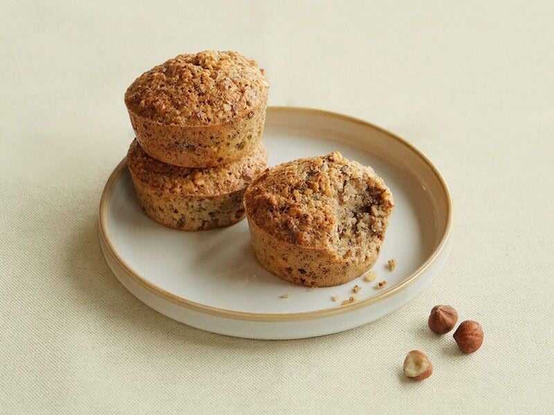 Trois muffins aux noisettes posés sur une assiette beige avec quelques noisettes à côté.