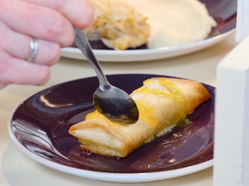Une main verse une sauce sur un dessert roulé doré posé sur une assiette violette.