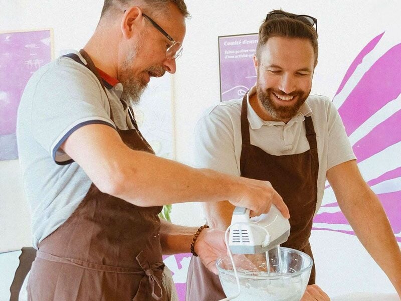 Deux hommes souriants battent une préparation dans un bol lors d'un cours de pâtisserie.