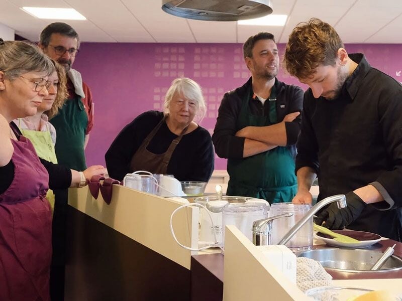 Un groupe d'adultes attentifs suit un cours de cuisine dans une salle équipée avec des plans de travail et des ustensiles.