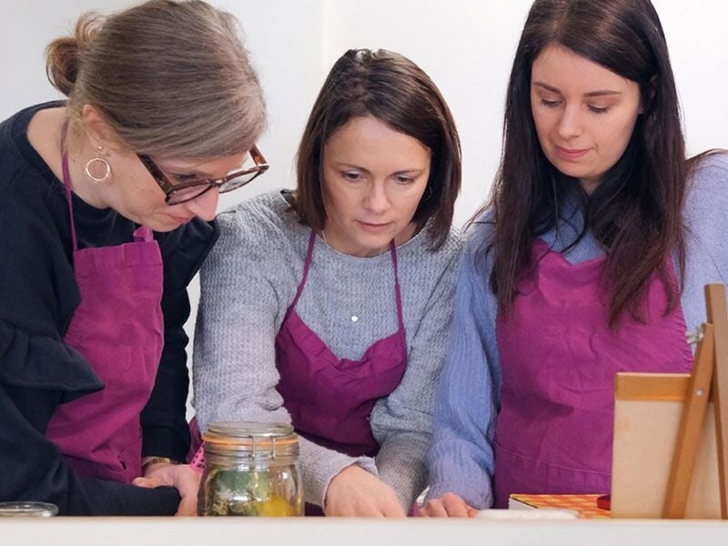 Trois femmes portant des tabliers roses observent attentivement un bocal lors d'un cours de cuisine en groupe.