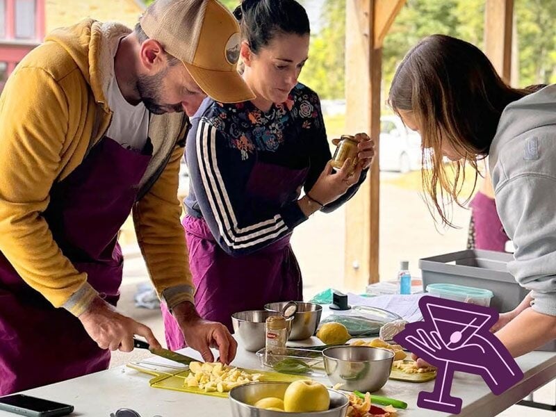 Trois personnes en tabliers préparent des ingrédients frais sur une table en extérieur lors d'un atelier culinaire.