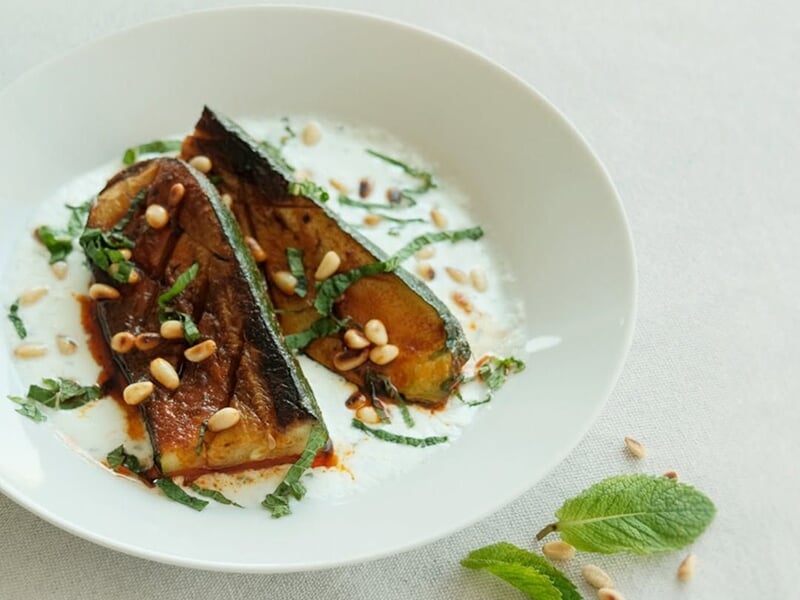 Un plat de poisson grillé avec une sauce blanche, des pignons et des herbes fraîches présenté dans une assiette blanche.
