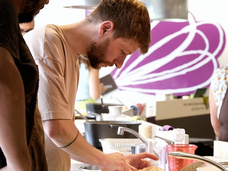 Un homme barbu en tablier beige prépare un plat avec concentration dans une cuisine équipée et colorée.