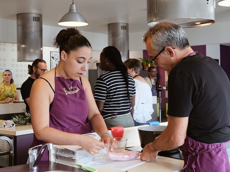 Deux personnes en tabliers préparent ensemble un plat dans une cuisine animée avec plusieurs participants.