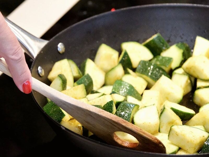 Une main remue des morceaux de courgettes dans une poêle noire avec une spatule en bois.