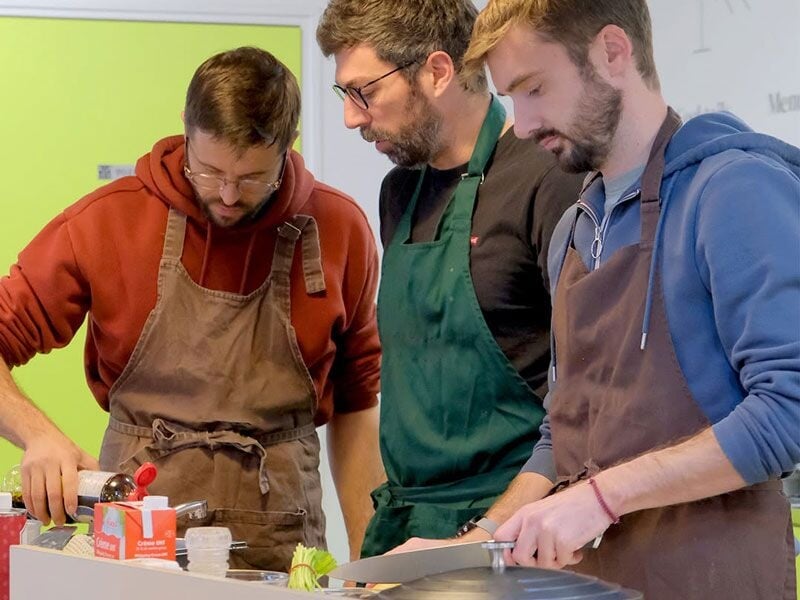 Trois hommes concentrés préparent des plats ensemble dans une cuisine équipée, portant des tabliers.