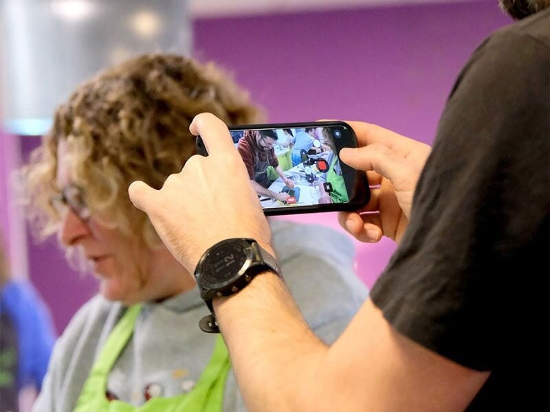 Une personne filme avec un smartphone un atelier de cuisine où plusieurs participants sont en pleine préparation.