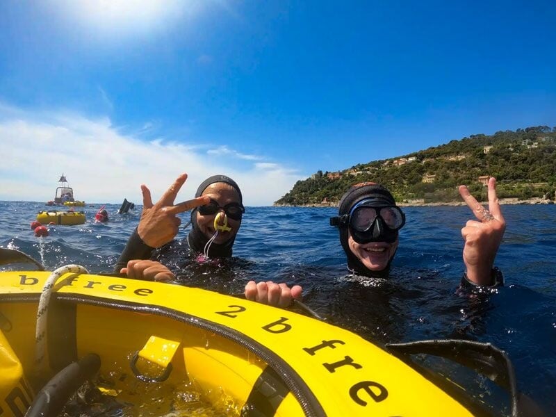 Deux plongeurs souriants en combinaison noire font un signe de paix près d'un bateau jaune en mer calme.