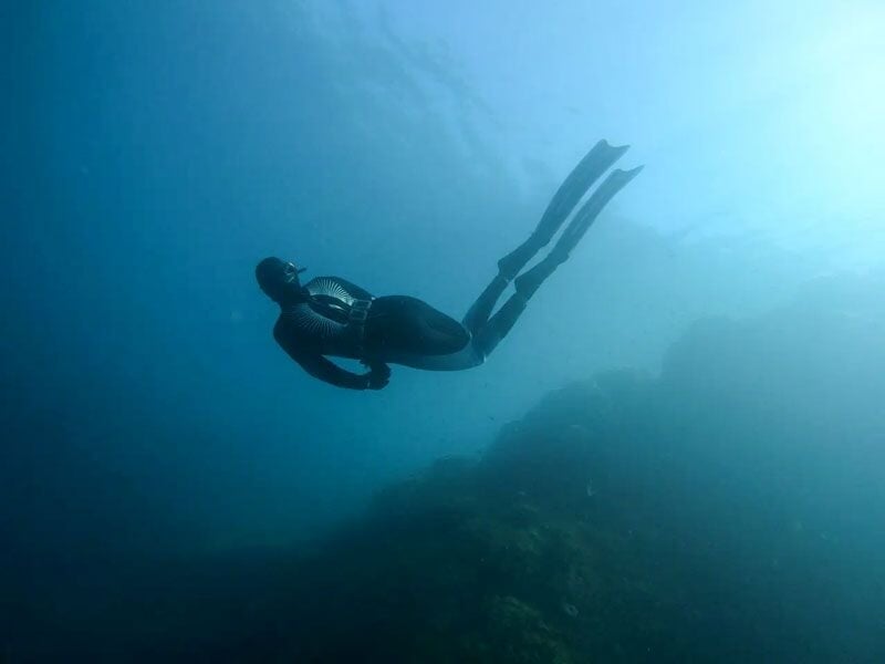 Plongeur en combinaison noire nageant sous l'eau au-dessus d'un fond rocheux dans une mer claire.