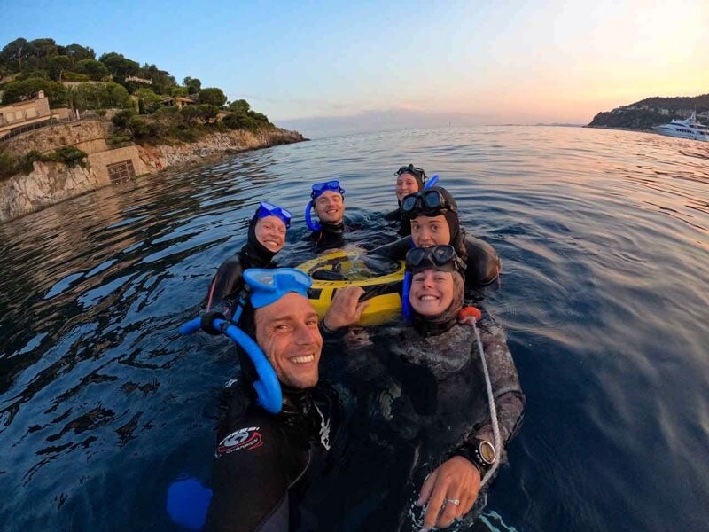 Groupe de cinq plongeurs souriants en surface au coucher du soleil, avec des masques et tubas bleus.