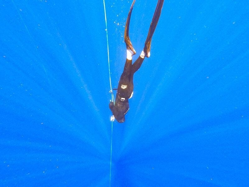Plongeur en combinaison noire suspendu verticalement sous l'eau, tenant une corde dans une mer très bleue.