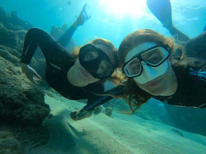 Deux plongeuses en masque et tuba nagent près du fond sableux et rocheux d'une mer claire.