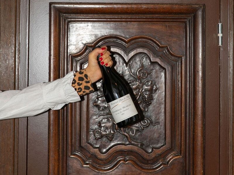 Main féminine tenant une bouteille de vin rouge devant une porte en bois sculpté, ambiance élégante.