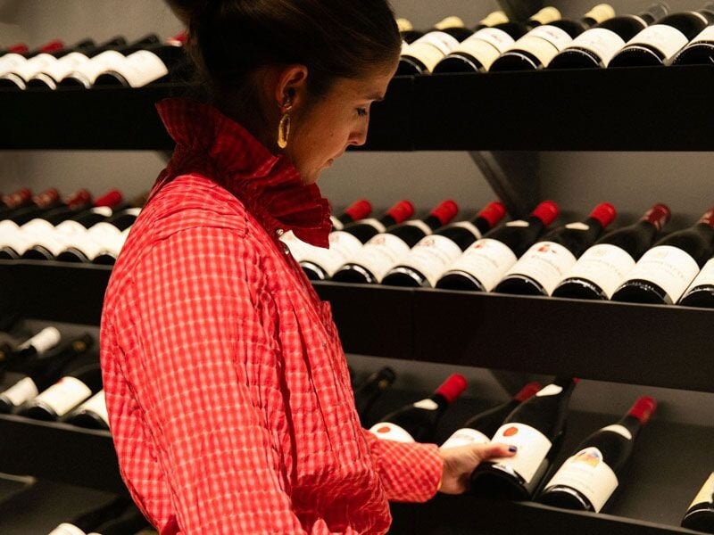 Femme en chemise rouge choisissant une bouteille de vin sur une étagère bien garnie dans une cave.