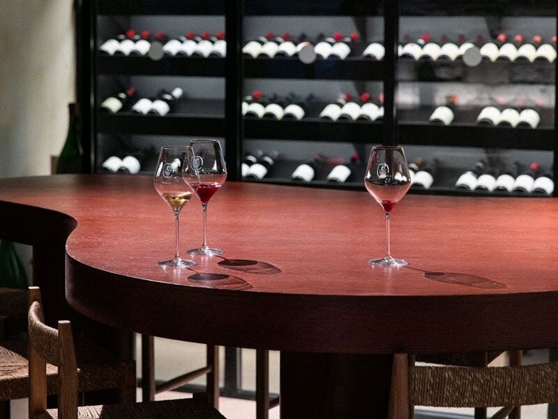 Trois verres de vin posés sur une table en bois, avec en arrière-plan des étagères remplies de bouteilles.