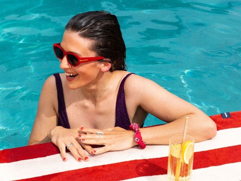 Femme souriante dans une piscine, portant des lunettes de soleil rouges et un cocktail à côté