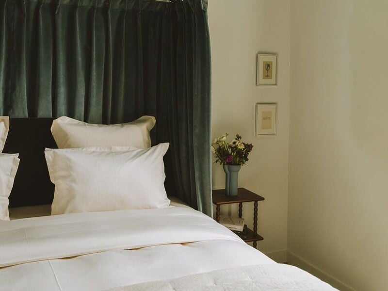 Chambre avec un grand lit blanc, tête de lit noire et rideaux verts foncés, table avec vase de fleurs