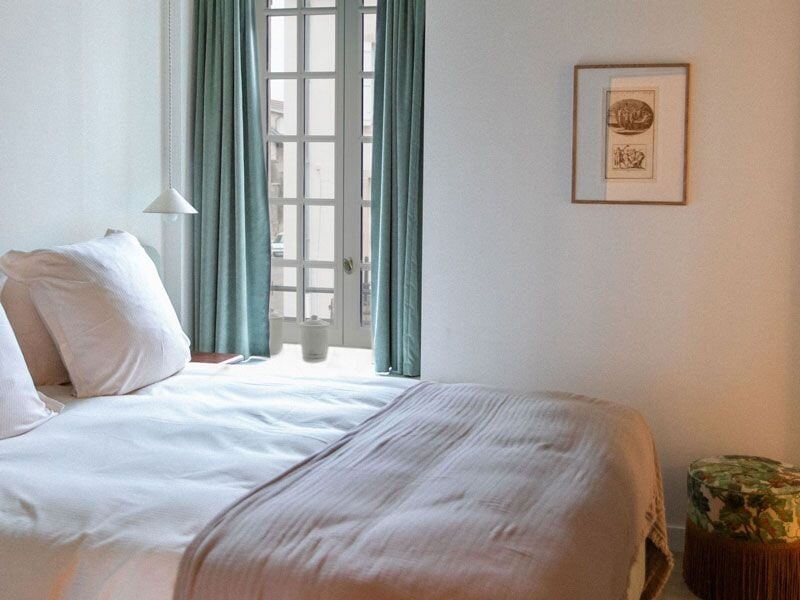 Chambre lumineuse avec lit blanc, couverture beige, rideaux verts et tableau encadré au mur