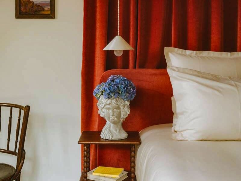 Intérieur d'une chambre avec rideaux rouges, lit blanc et vase décoratif en forme de tête avec fleurs bleues.