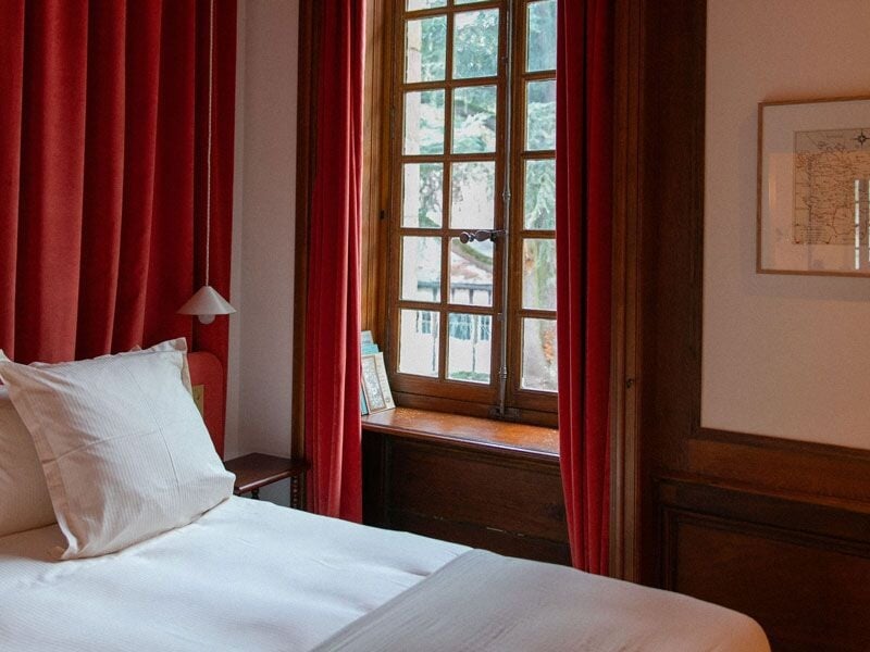 Chambre lumineuse avec lit blanc, rideaux rouges et grande fenêtre en bois laissant entrer la lumière naturelle.