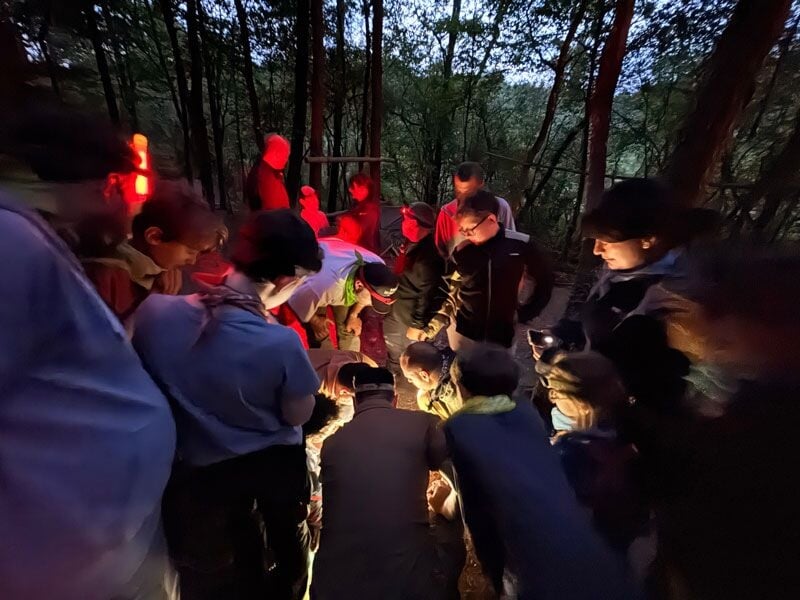 Groupe de personnes en forêt la nuit, rassemblées autour d'une source de lumière pour une activité de survie.