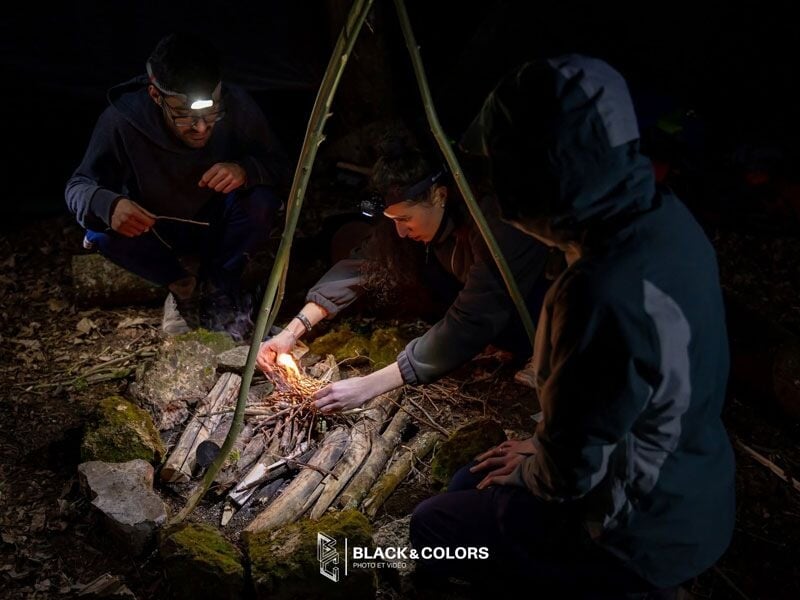 Trois personnes en pleine forêt la nuit, allumant un feu de camp avec des brindilles sous un abri improvisé.