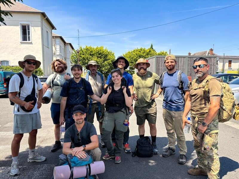 Groupe de neuf personnes prêtes pour un stage de survie, équipées de sacs à dos et vêtements adaptés en extérieur.