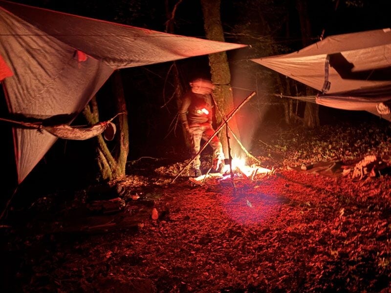 Campement de nuit en forêt avec un feu de camp allumé et des bâches tendues pour abriter les participants.