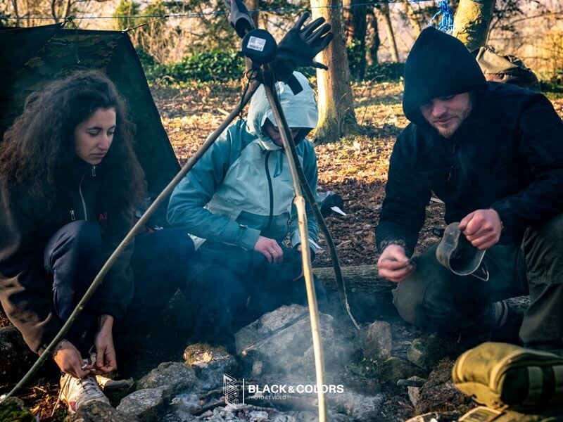Trois personnes autour d'un feu de camp en forêt, préparant ou cuisinant quelque chose dans une ambiance conviviale.