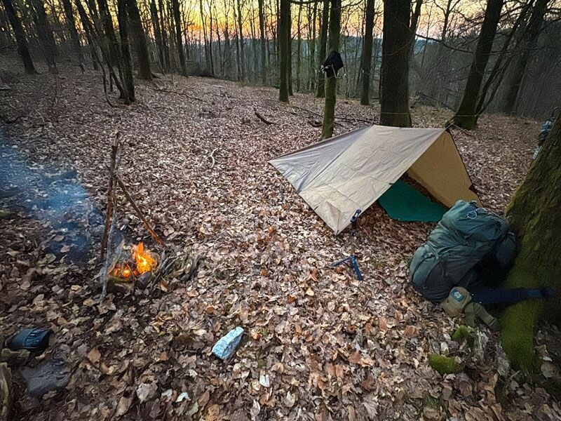 Petit campement en forêt avec une tente de fortune, un sac à dos et un feu de camp fumant au sol.