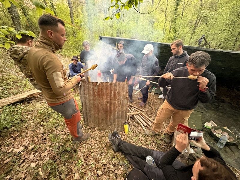 Groupe de personnes en forêt autour d'un feu, certains cuisinent tandis qu'un autre filme la scène.