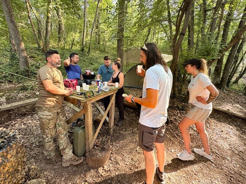Groupe de personnes en forêt discutant autour d'une table en bois, dans une ambiance détendue et conviviale.