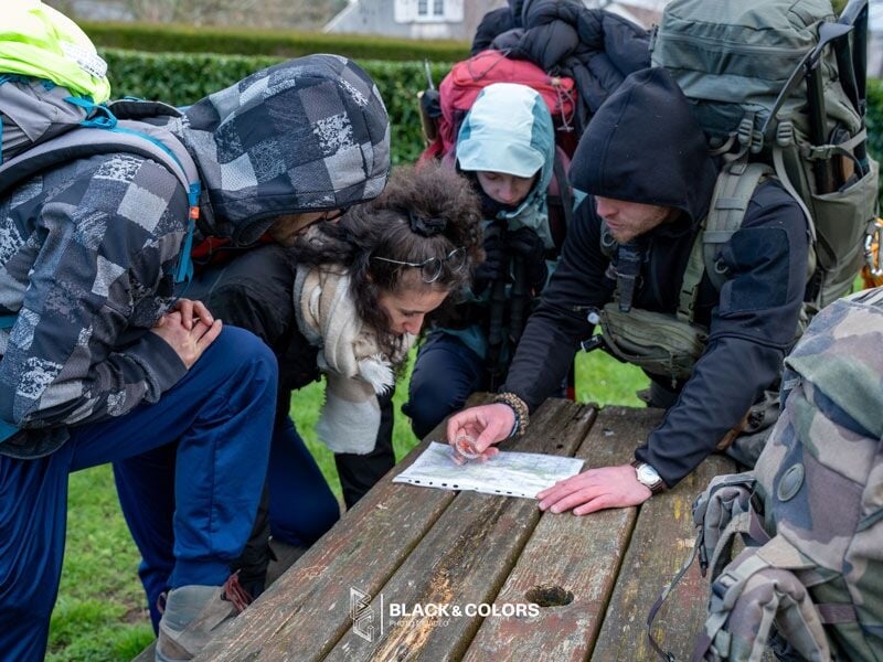 Quatre randonneurs en tenue chaude consultent une carte sur une table en bois dans un parc vert.