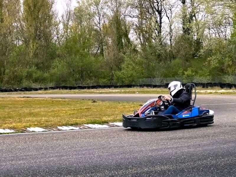 Personne en casque blanc conduisant un kart bleu sur une piste de karting entourée d'herbe et d'arbres verts.