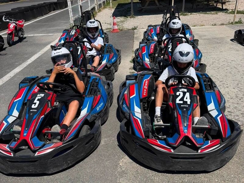 Quatre jeunes en karts bleus et rouges portant des casques blancs, prêts à démarrer sur une aire de karting.