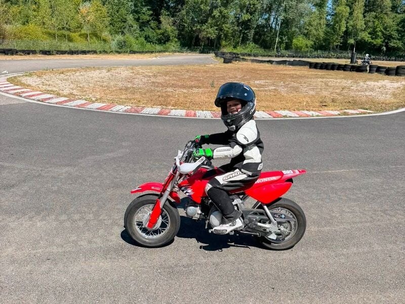 Enfant en tenue de moto et casque noir, roulant sur une petite moto rouge sur circuit fermé en plein jour.