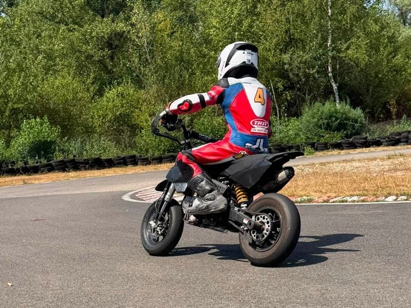 Jeune pilote en combinaison rouge et bleue avec casque blanc, sur une moto noire, sur circuit entouré d'arbres.