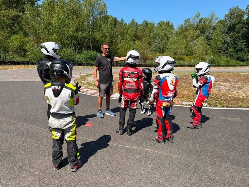 Groupe d'enfants en combinaisons de moto et casques, écoutant un instructeur sur un circuit de pilotage moto.