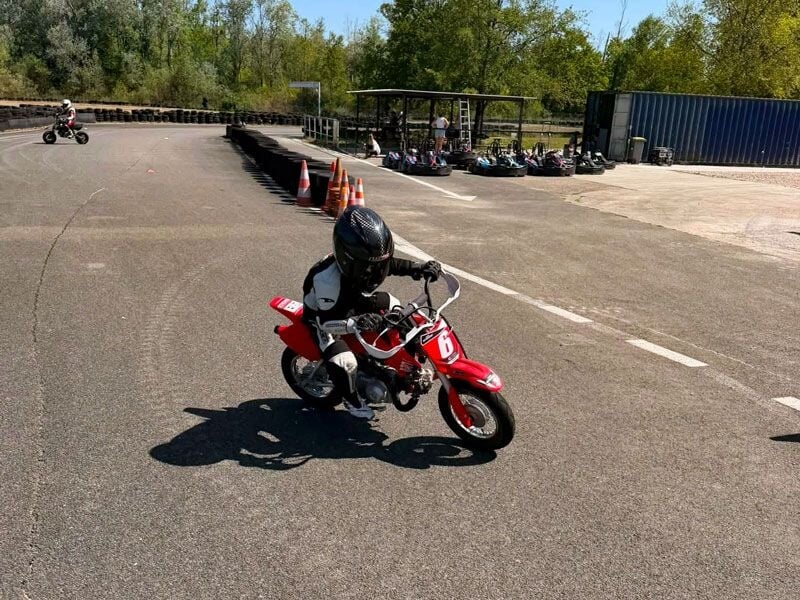 Jeune pilote en tenue noire et casque noir, prenant un virage sur une petite moto rouge sur circuit asphalté.